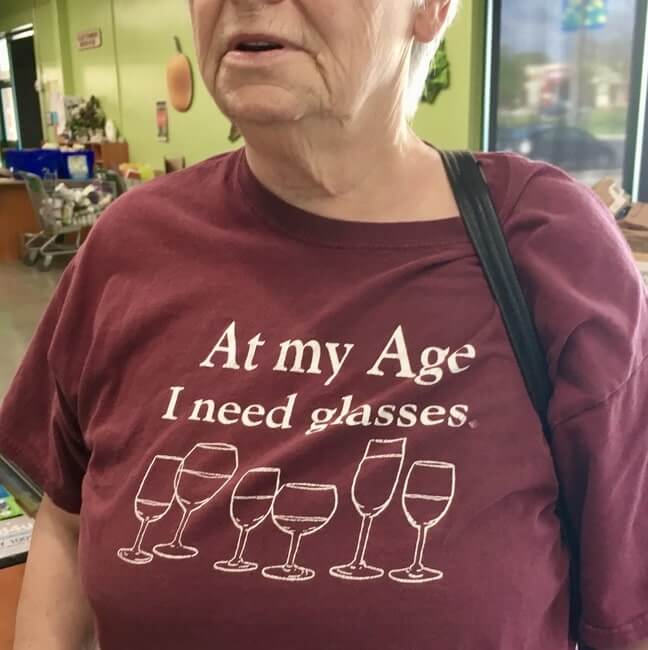 Older woman wearing t-shirt with image of wine glasses that reads: "At my age I need glasses"